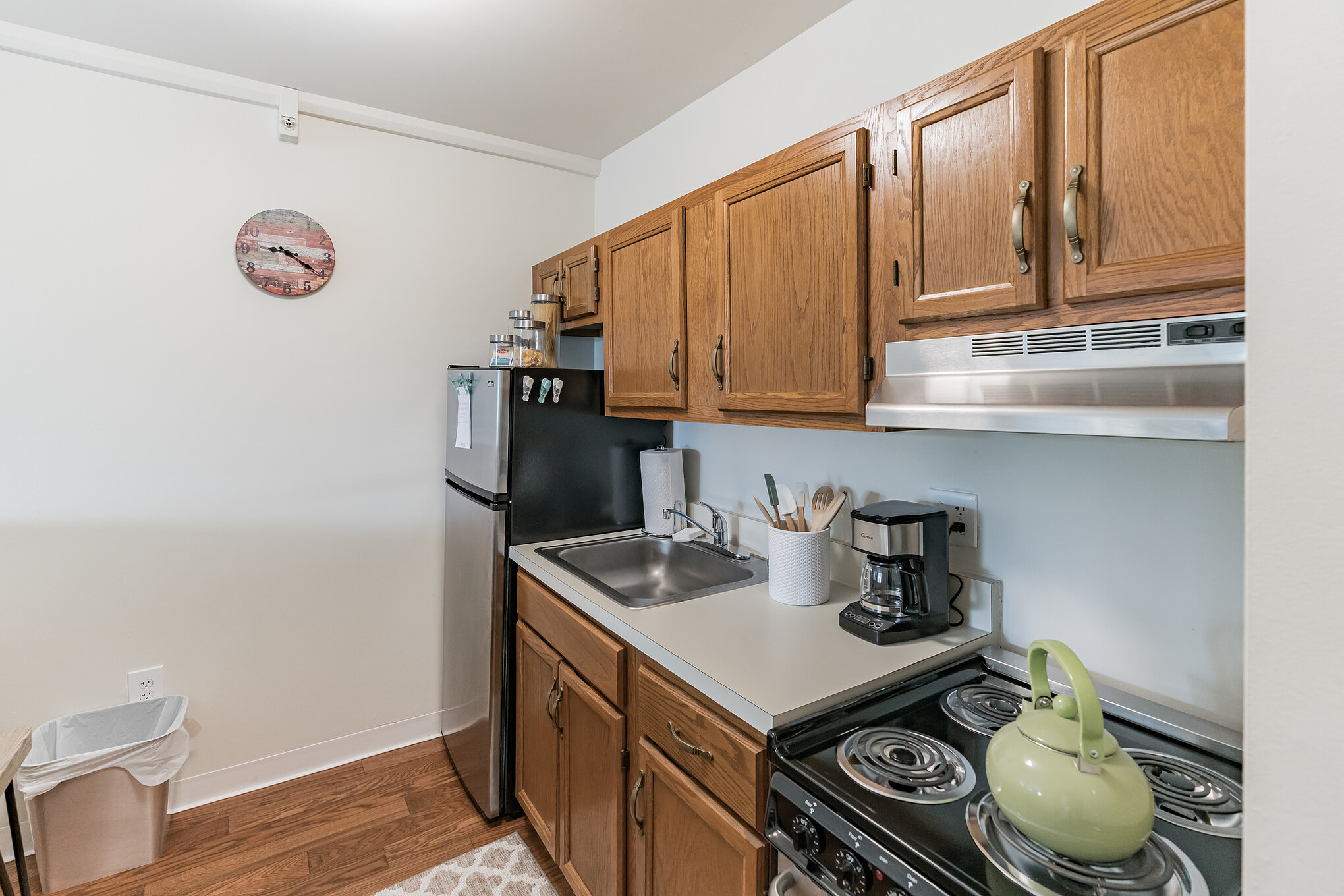 14560-STD-3-11205046 - Shorehaven A kitchen with an American flag-patterned circular wall clock, light-brown cabinets, a fridge, sink, a coffee maker, and a green kettle on the gas stove at Shorehaven, a top choice for senior living near Warren.