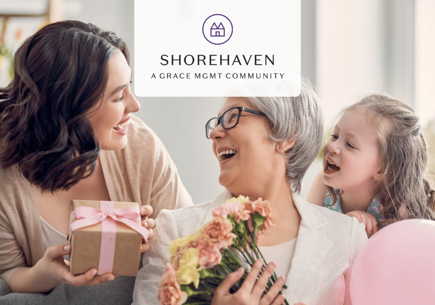 Young woman hands a wrapped gift to an elderly woman who holds a bouquet, with a smiling child beside them in a bright room.