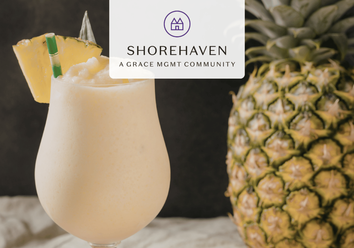 Cream-colored tropical cocktail in a curved glass with a pineapple wedge and green straw, foregrounded with a whole pineapple and Shorehaven branding overlay on top.